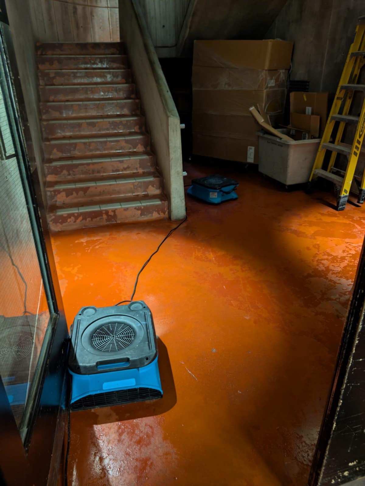 Standing water across a commercial floor at the foot of a stairwell with an air mover running