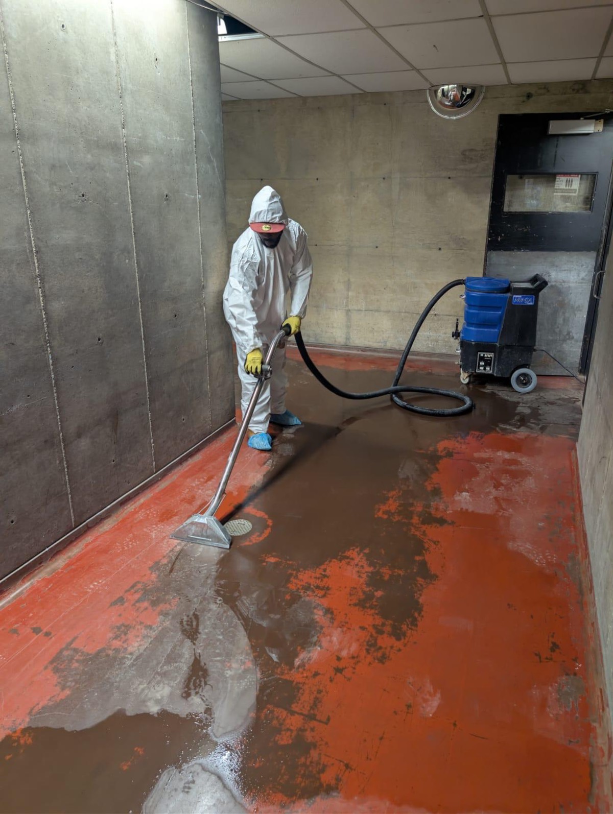 Technician in a protective suit and respirator pulling standing water off a concrete floor with an extraction wand